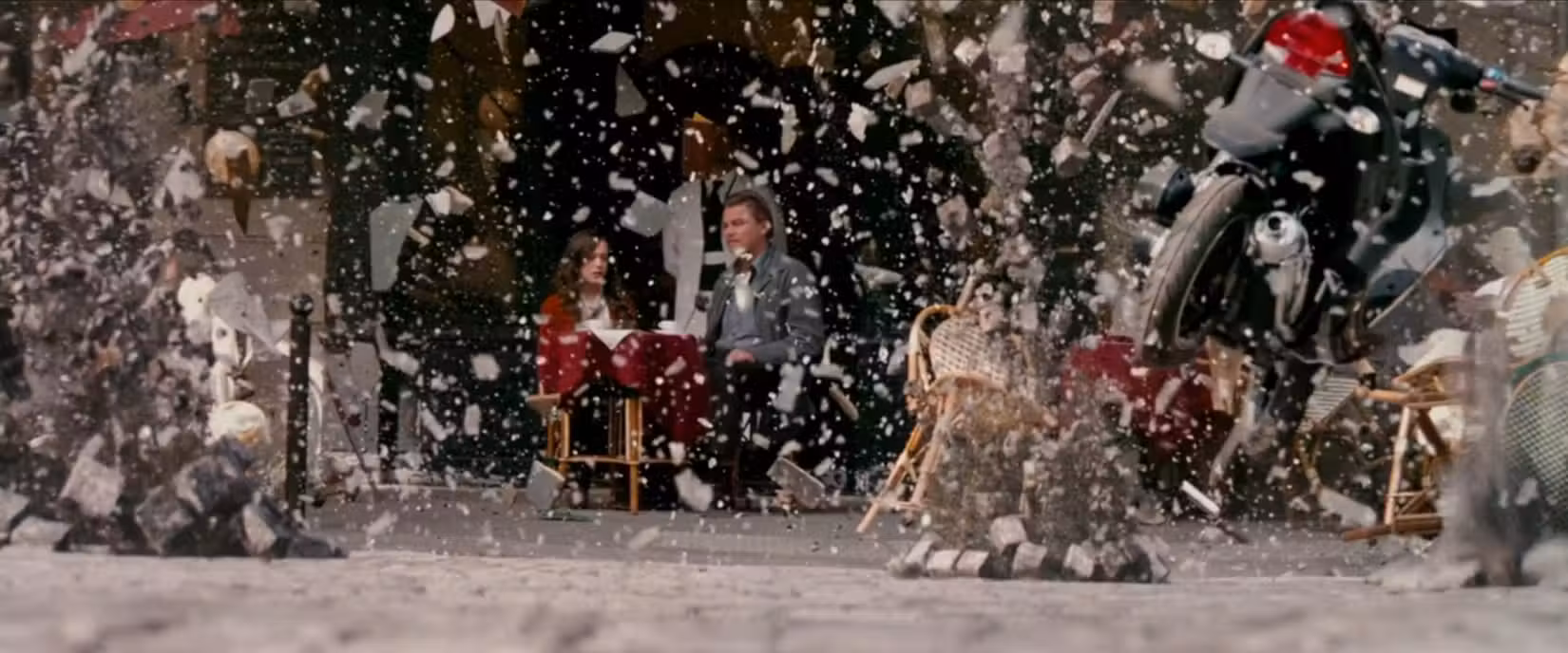 Dom (Leonardo DiCaprio) e Ariadne (Elliot Page) sentam-se em um café, obscurecidos por objetos explodindo ao seu redor enquanto um ambiente de sonho se desfaz em uma cena do filme A Origem, de Christopher Nolan.