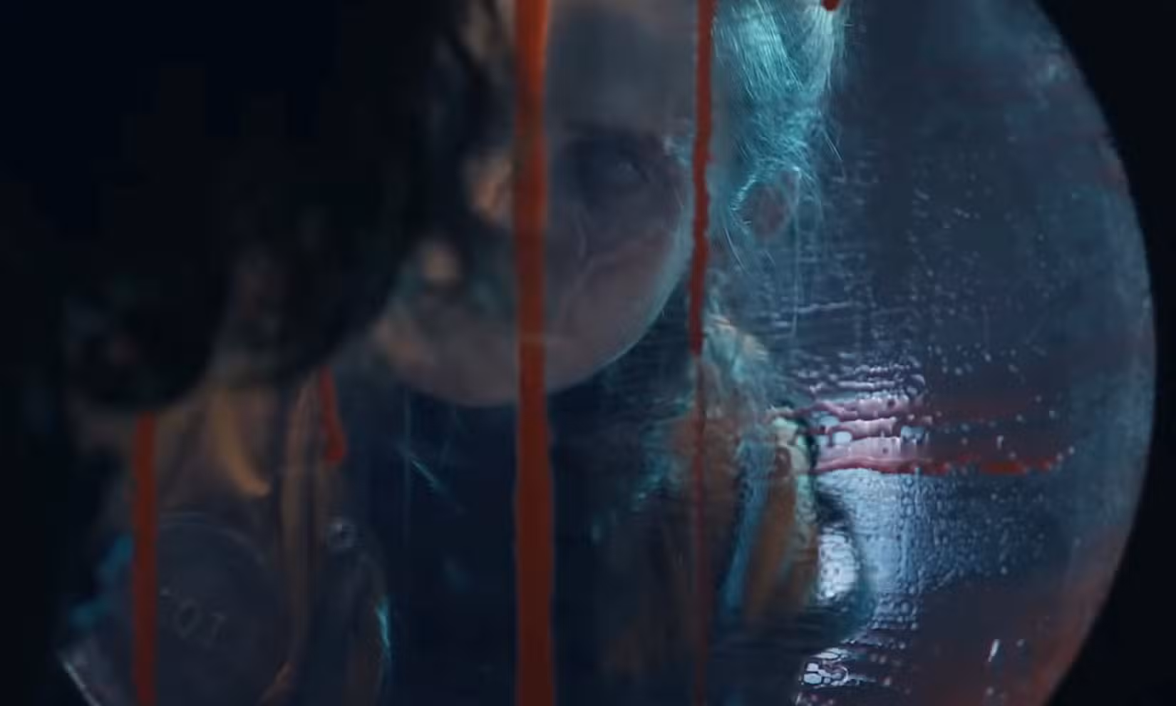 Uma mulher com um olho completamente branco e uma cicatriz que corta uma bochecha olha através de uma vigia pingando sangue no Pulmão de Ferro