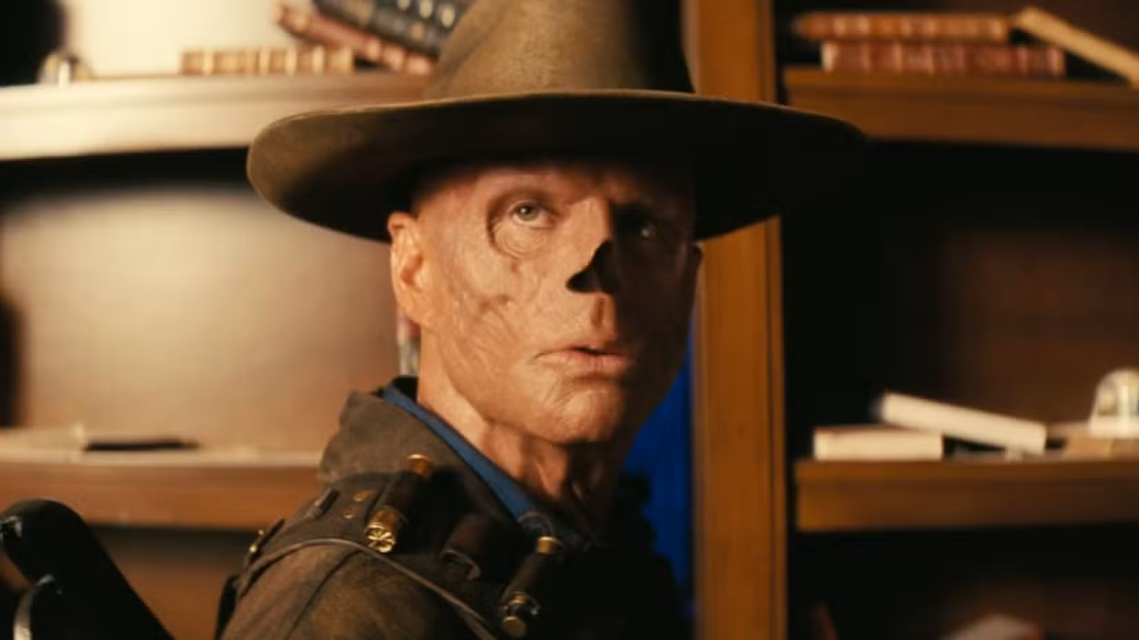 Uma imagem de Walton Goggins como o Ghoul da 2ª temporada de Fallout. Sua pele é irregular e ele não tem nariz. Ele usa um chapéu stetson e um pó de couro. Ele se vira para alguém fora da tela, parecendo irritado.
