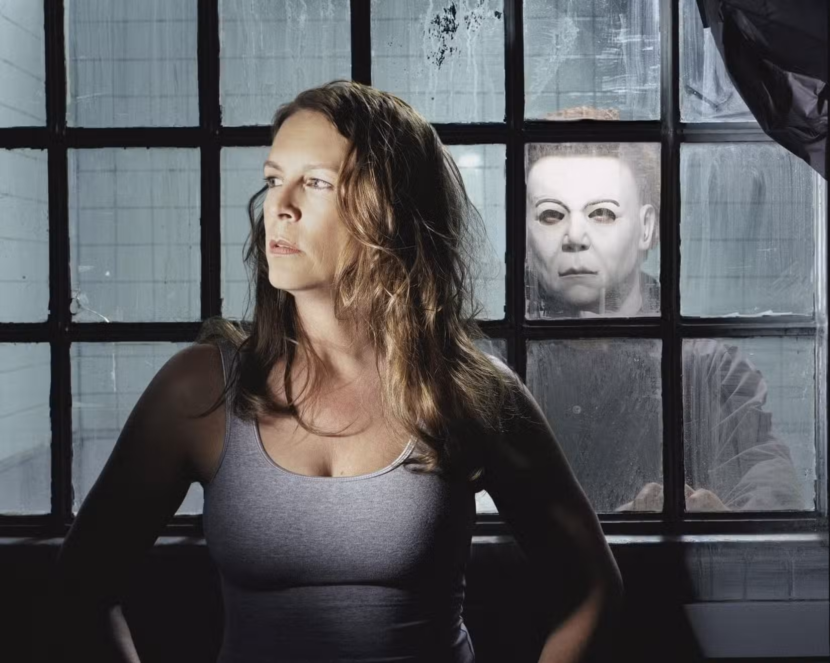 Laurie Strode (Jamie Lee Curtis) está em frente a uma série de janelas, com o mascarado Michael Myers esperando do outro lado, em cena de Halloween: Ressurreição (2002).