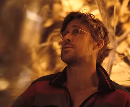 O astronauta Ryland Grace (Ryan Gosling), de cabelos desgrenhados, usando óculos e macacão laranja, diante de um fundo facetado de cristal