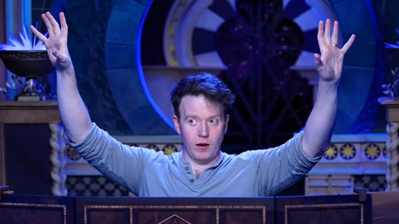 Uma imagem de Brennan Lee Mulligan atrás de uma tela do Dungeon Master. Atrás dele está um fundo de fantasia, inundado de luz azul.