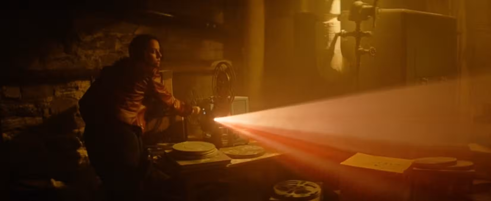 Claire (Kaya Scodelario) liga um projetor de filme antigo, transmitindo uma luz vermelha por uma sala iluminada de amarelo, em uma cena de Resident Evil: Bem-vindo a Raccoon City