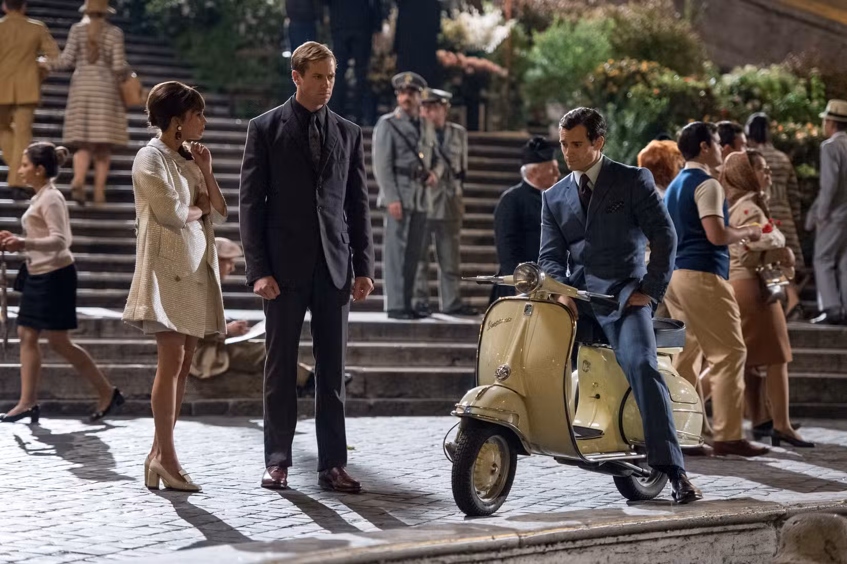 Em uma cena média do filme The Man from UNCLE, de Guy Ritchie, Henry Cavill está sentado em uma scooter ao lado de Armie Hammer e Alicia Vikander em pé, todos vestidos com roupas retrô dos anos 60