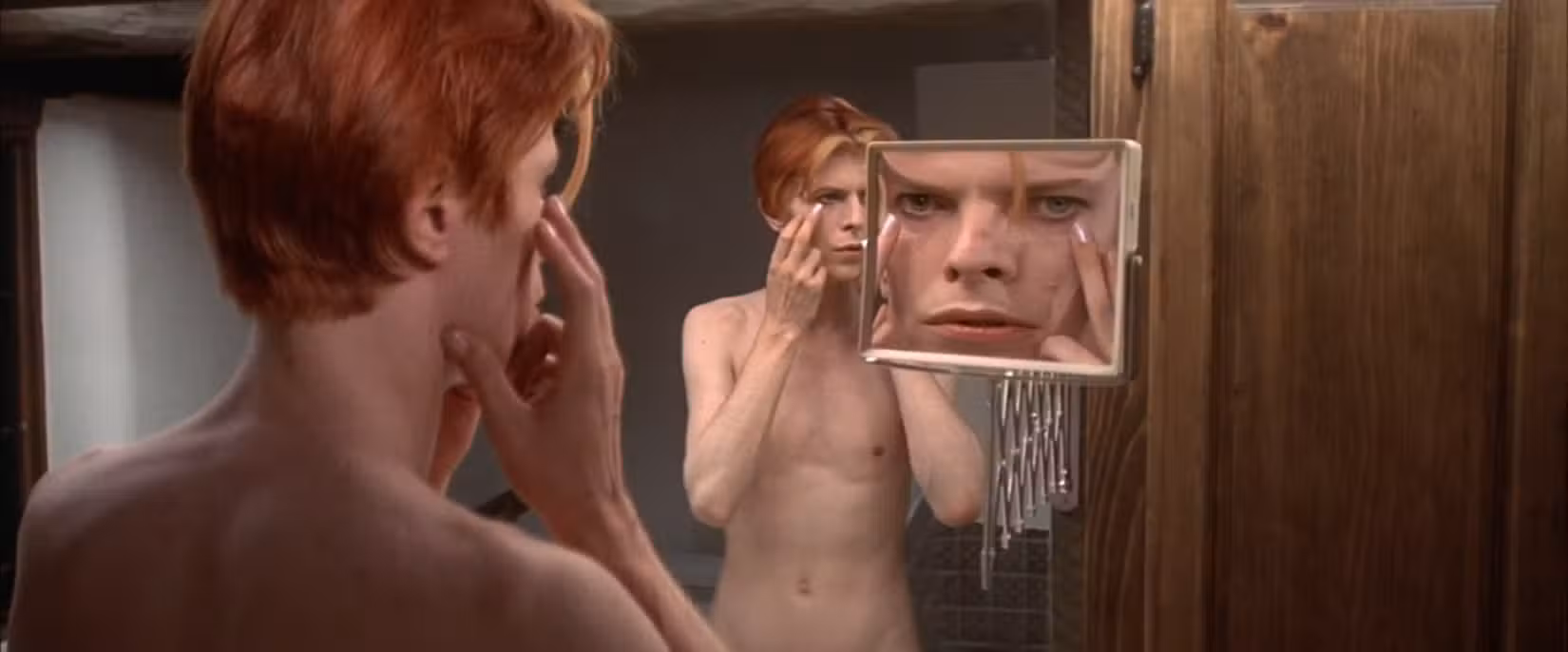 Um alienígena nu (David Bowie) examina seu rosto humano em uma série de espelhos, um dos quais distorce seu rosto em close-up, em cena de O Homem que Caiu na Terra (1976).