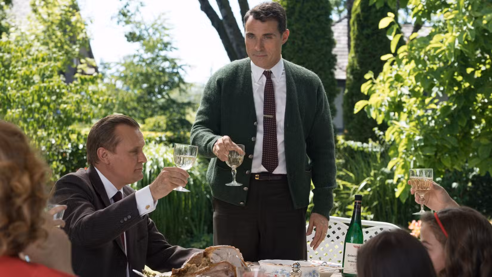 John Smith (Rufus Sewell) segura uma taça de vinho para um brinde em uma festa no jardim em O Homem do Castelo Alto