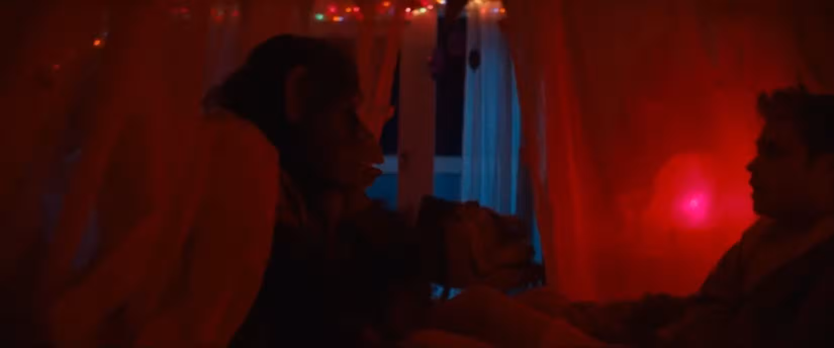 Um chimpanzé raivoso está sentado na beira de uma cama, ameaçando um jovem irmão, todo mostrado de perfil e banhado por luz vermelha em uma cena de Primata.