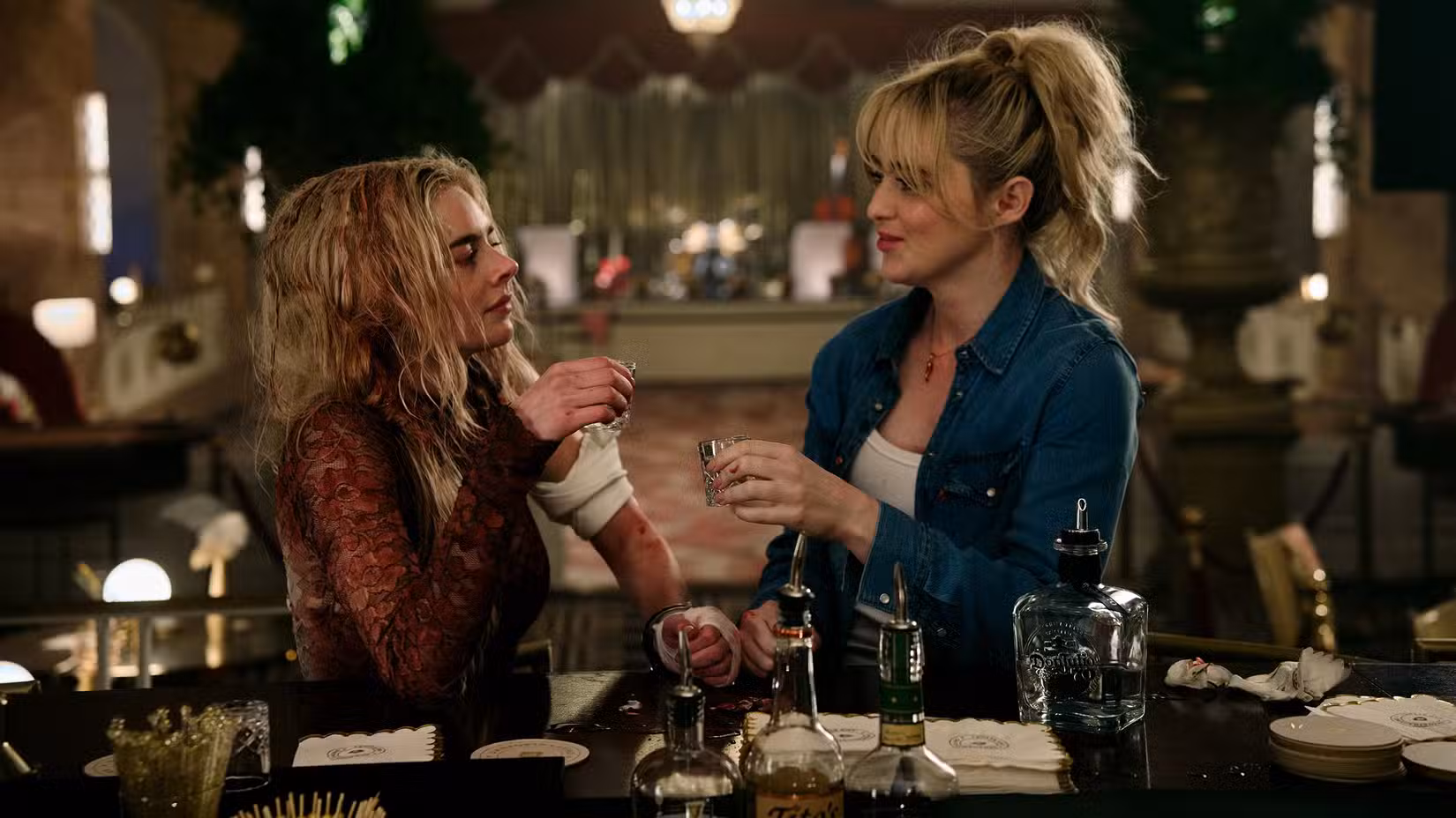 Grace (Samara Weaving), uma mulher usando um vestido de noiva encharcado de sangue, e Faith (Kathryn Newton) vestindo uma camiseta jeans, compartilham doses de tequila no salão de baile de um hotel.