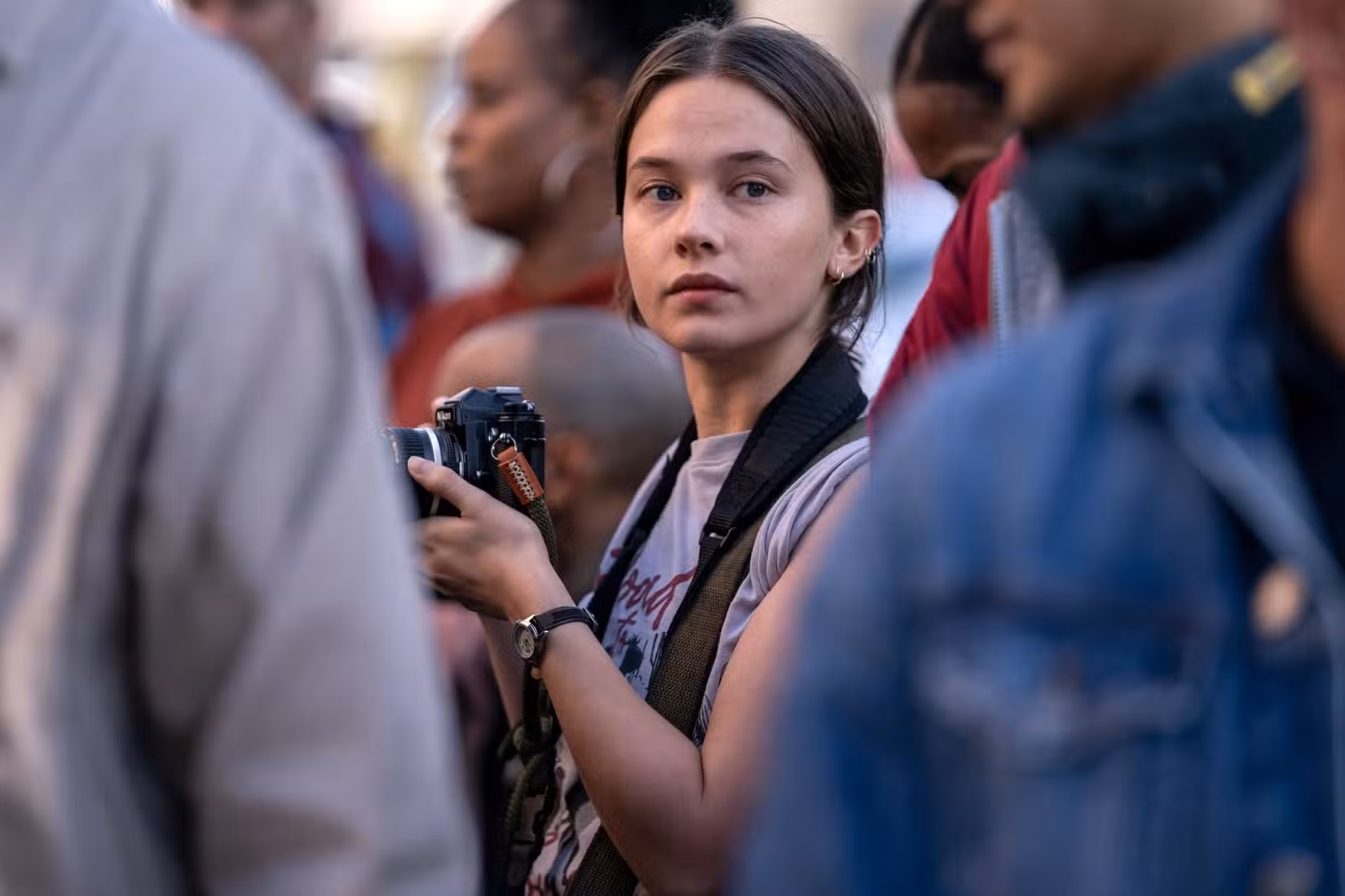 Jessie (Cailee Spaeny), uma jovem fotojornalista segurando uma câmera, está no meio de uma multidão em Guerra Civil, de Alex Garland.