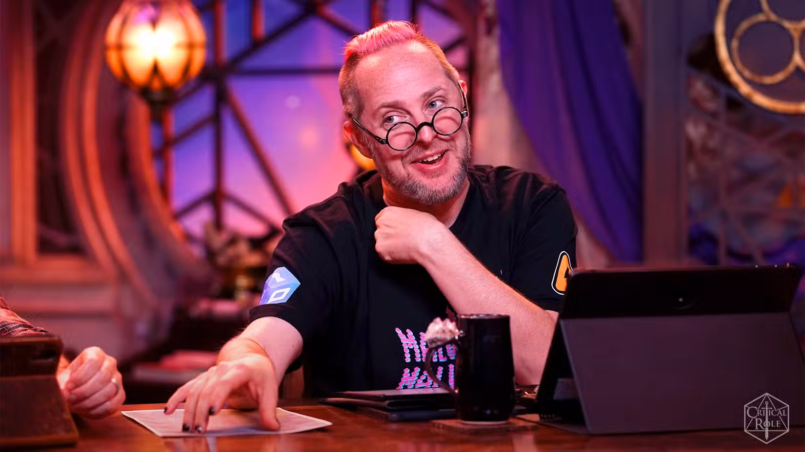 Uma imagem de Taliesin Jaffe em Critical Role. Ele está sentado à mesa, com a mão sobre um pedaço de papel. O outro está em seu pescoço. Ele está falando.