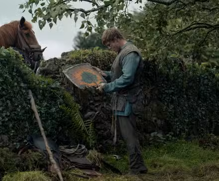 Dunk (Peter Claffey), um homem alto e maltratado em roupas medievais, segura um escudo de madeira quebrado e o examina. Ele está parado em frente a uma cerca viva com um cavalo espiando por cima dela. De Um Cavaleiro dos Sete Reinos.