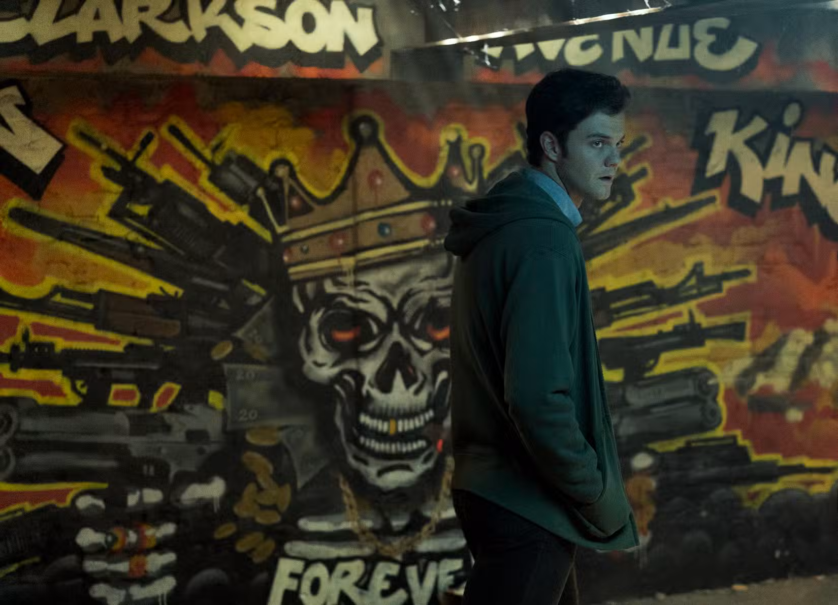 Jack Quaid como Hughie em The Boys, passando em frente a um mural de uma caveira coroada e envolta em armas