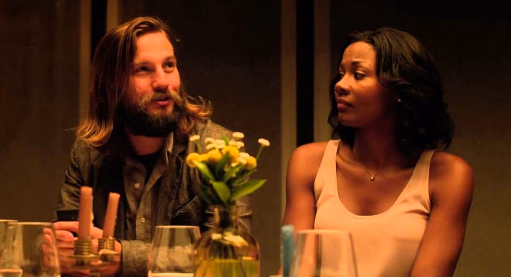 Um casal (Logan Marshall-Green e Emayatzy Corinealdi) está sentado lado a lado em uma mesa de jantar chique, com ela olhando para ele com cautela, em cena de O Convite (2016).