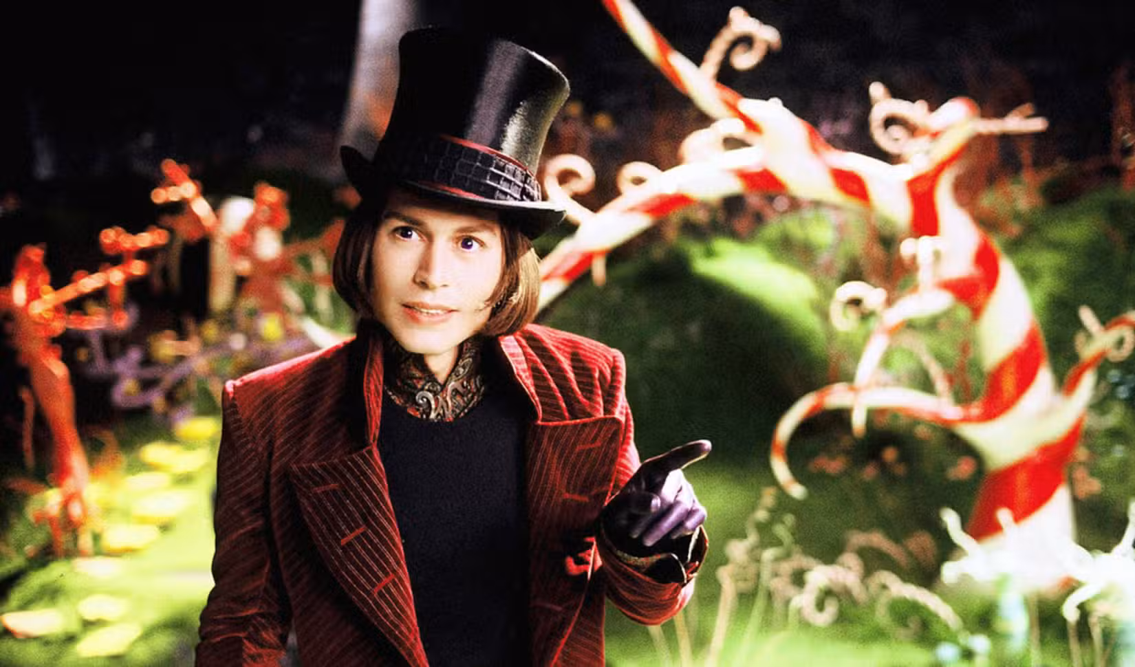 Willy Wonka (Johnny Depp, de paletó de veludo vermelho e cartola preta brilhante) sorri diante de um jardim feito de doces em Charlie e a Fábrica de Chocolate, de Tim Burton, de 2005.