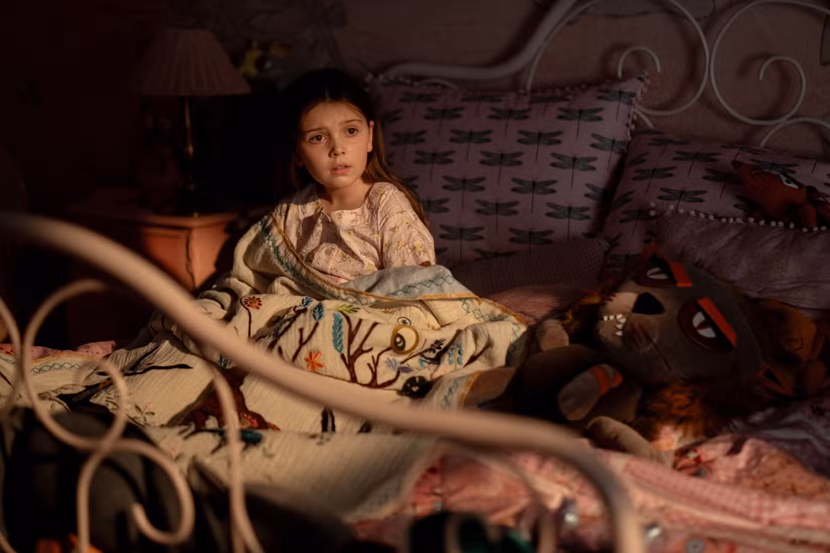 Uma imagem de Dust Bunny de Bryan Fuller apresentando Sophie Sloan como Aurora. Aurora está sentada de pijama na cama, cercada por seus brinquedos e travesseiros.