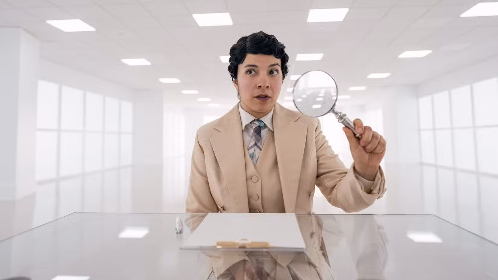 Muriel (Quelin Sepulveda), um anjo vestindo um terno bege e segurando uma lupa, está sentado em uma sala totalmente branca no céu em Good Opens.