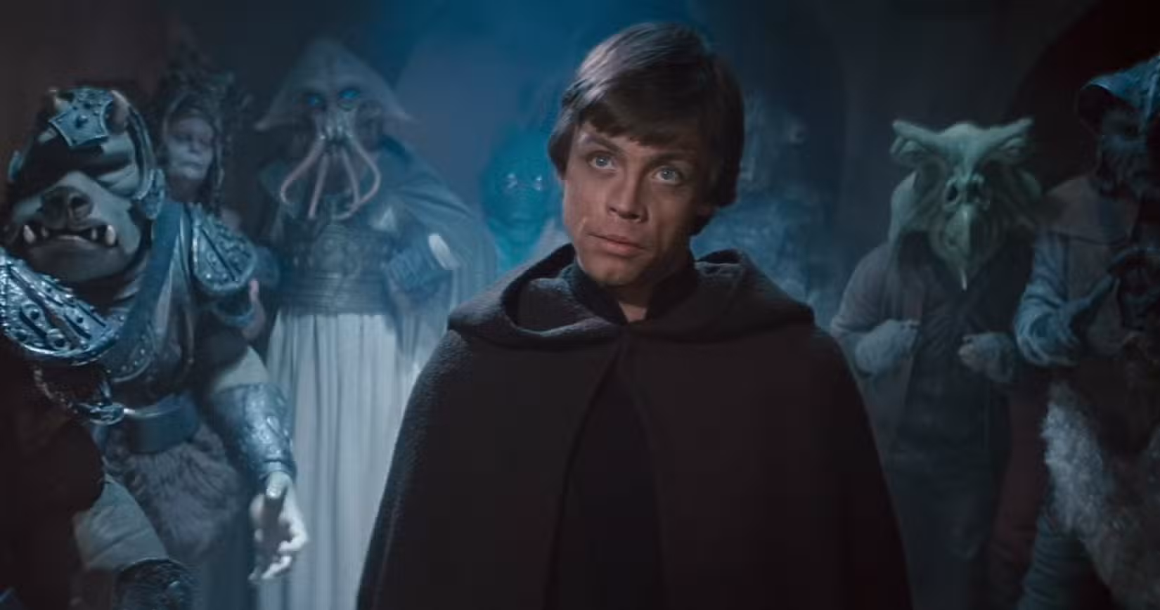 Luke Skywalker está no Palácio de Jabba, vestindo uma capa preta com capuz, com uma multidão de não-humanos atrás dele em Star Wars: O Retorno de Jedi