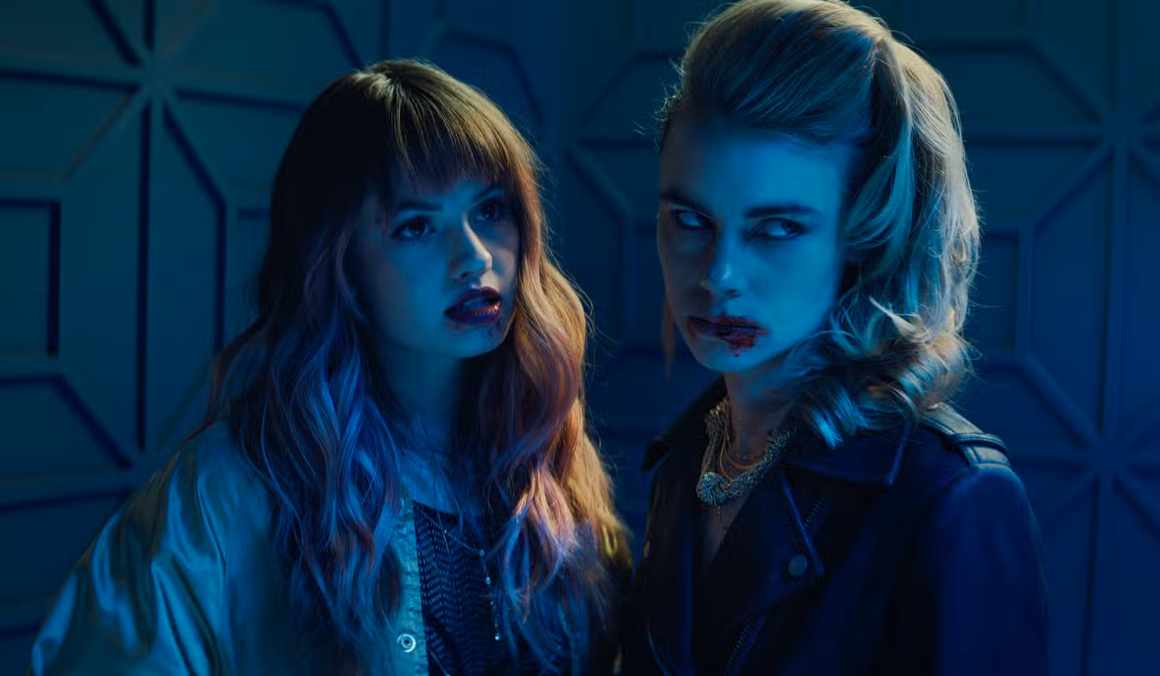 Dois dos vampiros de Night Teeth, Blaire (Debby Ryan) e Zoe (Lucy Fry), estão juntos em uma sala mal iluminada, com a boca ensanguentada.