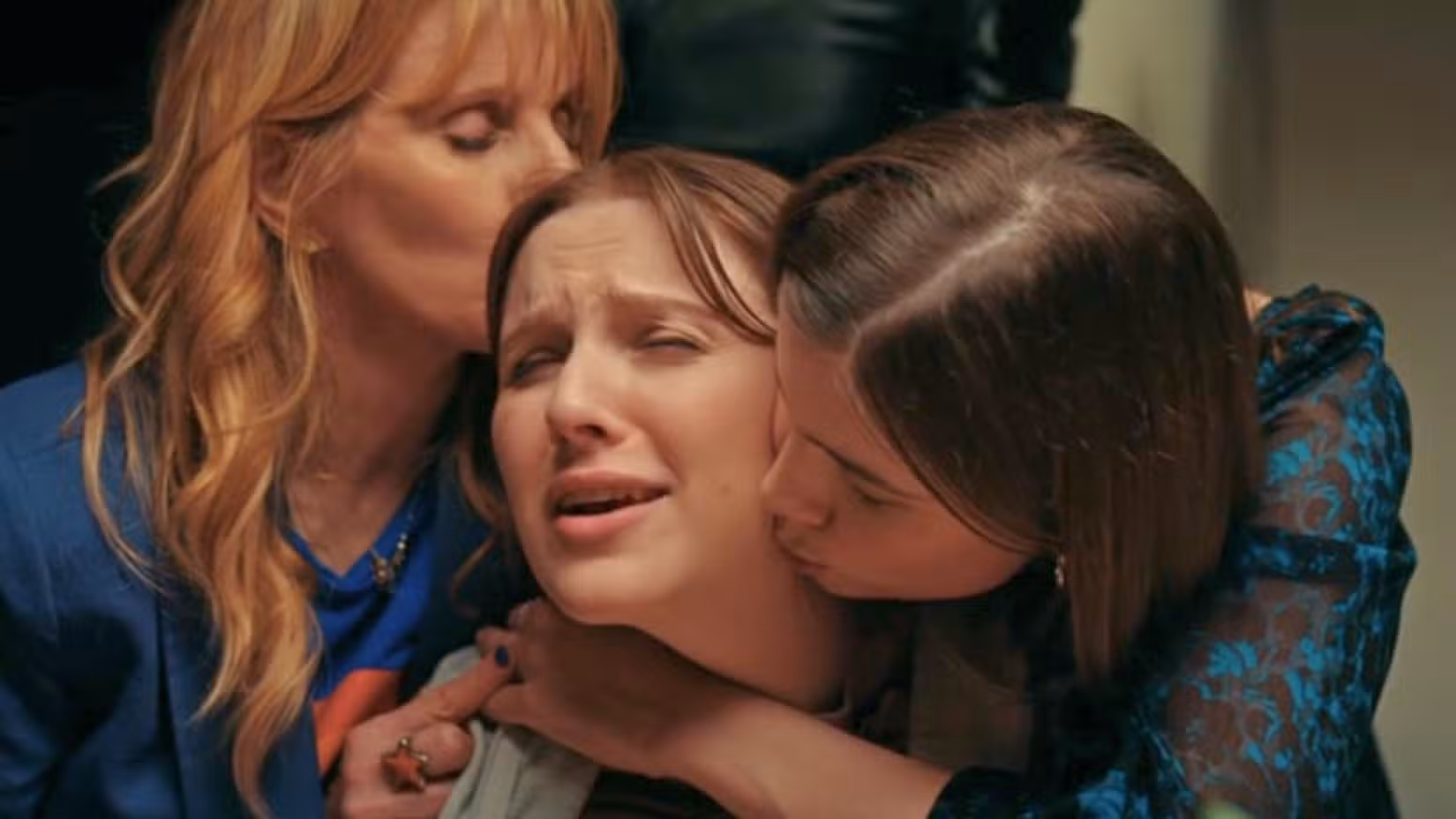 Uma imagem de D(e)ad. Mostra uma senhora loira mais velha beijando a cabeça da filha, que também está sendo abraçada pela irmã. A filha do meio parece irritada.