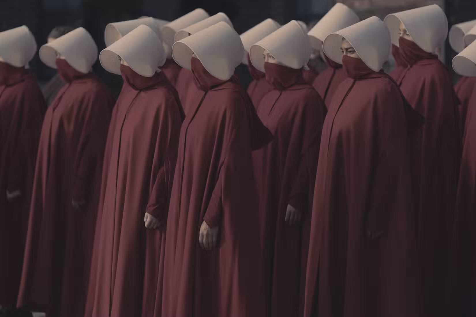 Uma fileira de aias em The Handmaid's Tale, mulheres com túnicas vermelho-escuras, máscaras faciais e toucas brancas, parecendo sóbrias