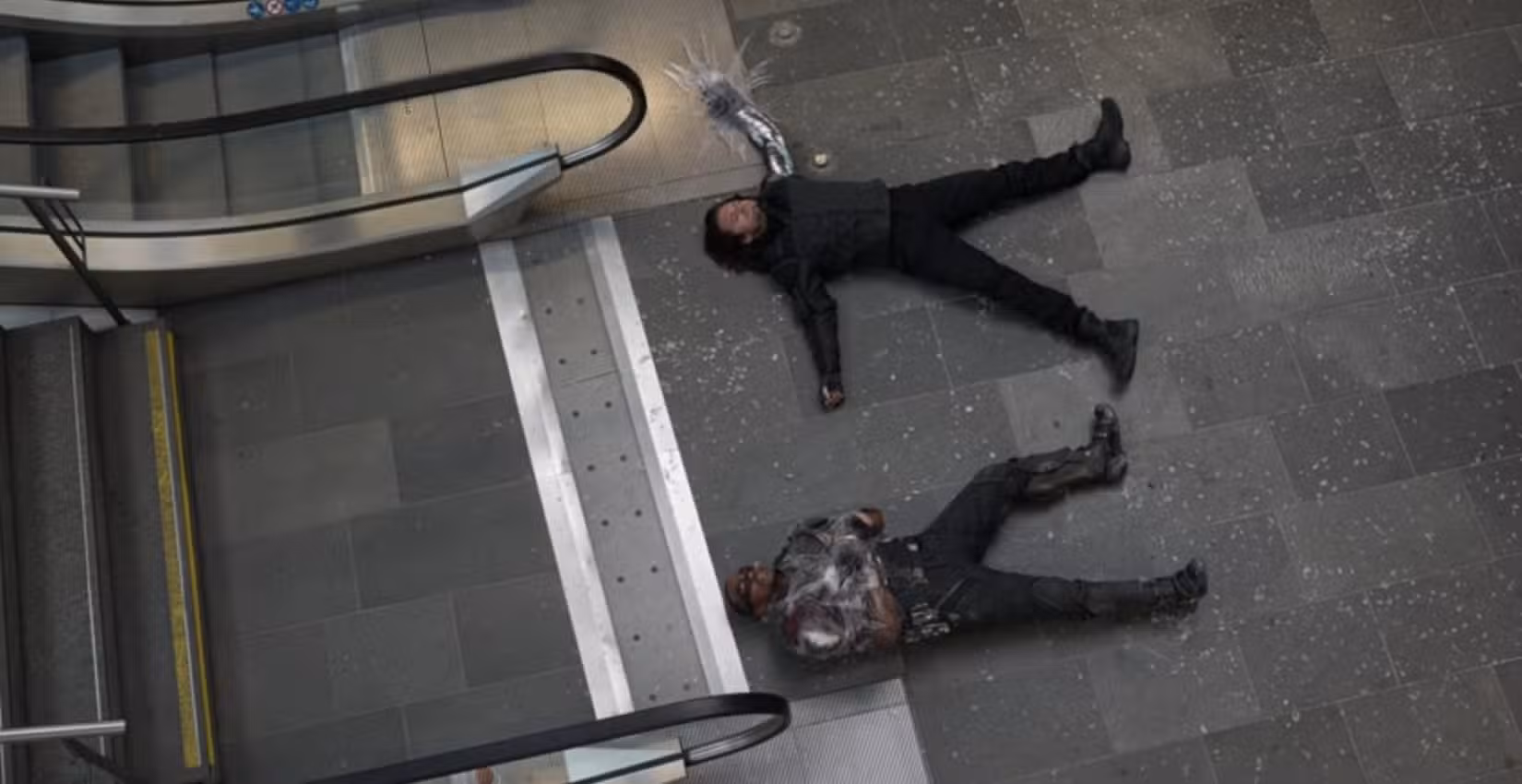 Em uma tomada aérea dessaturada de Capitão América: Guerra Civil, Bucky (Sebastian Stan) e o Falon (Anthony Mackie) estão derrotados no chão de concreto cinza, vestindo trajes acinzentados e azul marinho