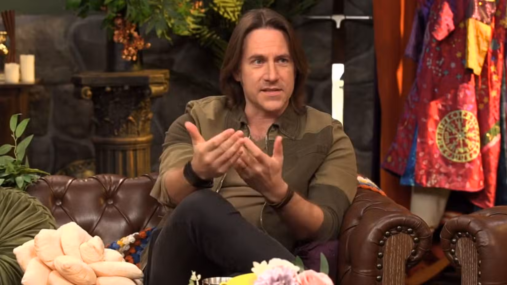 Uma imagem que mostra Matthew Mercer sentado em um sofá. Ele veste uma jaqueta verde e marrom e seu cabelo cai acima dos ombros. Ele move as mãos enquanto explica algo.