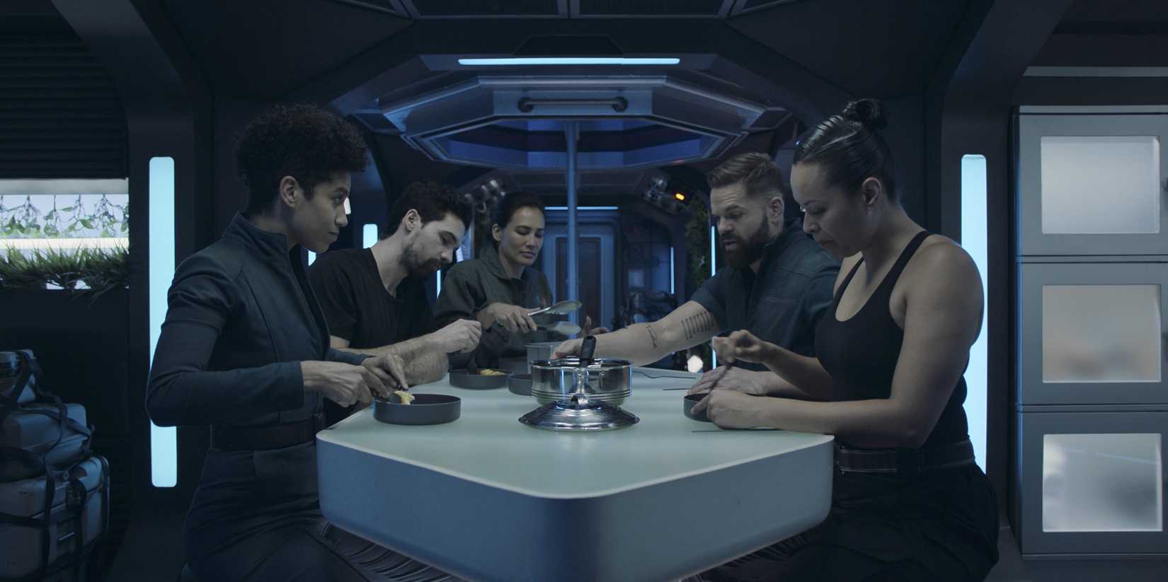 Cinco tripulantes de uma nave espacial se reúnem em torno de uma mesa futurística sob iluminação azul na 6ª temporada de The Expanse