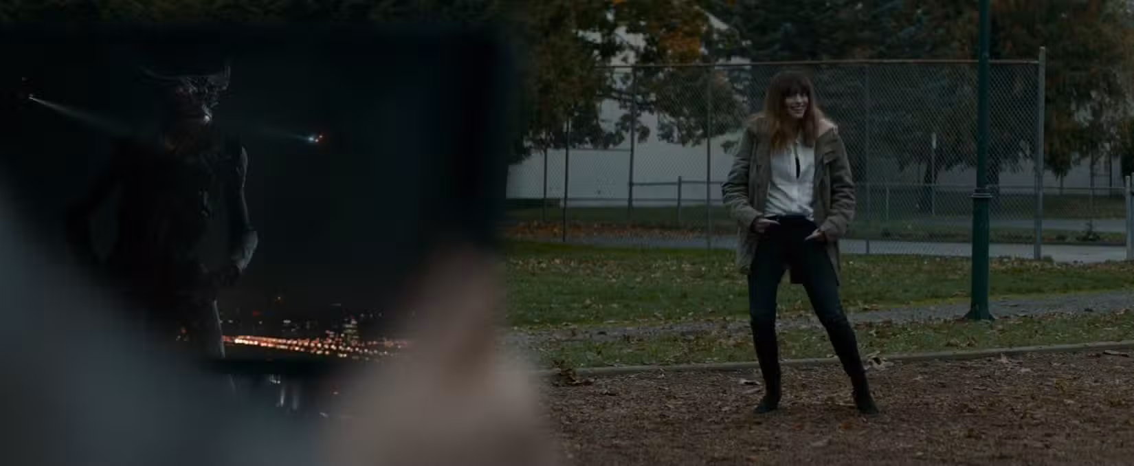 Em uma cena de Colossal (2017), Gloria (Anne Hathaway) está em um parque, em foco, mas no fundo da imagem, posando em uníssono com uma criatura kaiju vista em primeiro plano da imagem em um tablet, estabelecendo sua conexão bizarra.