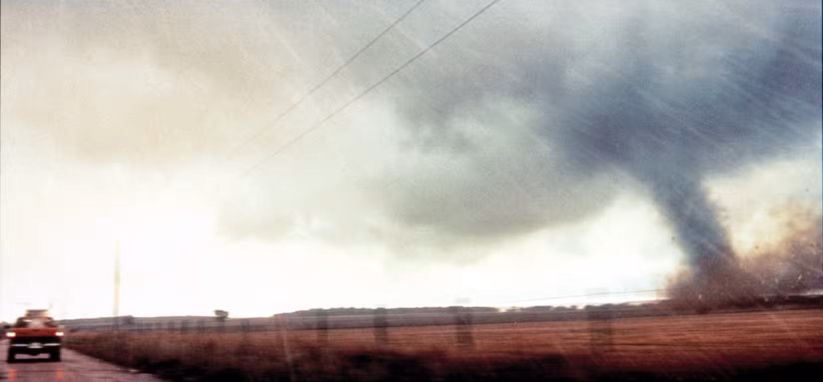 Uma imagem widescreen de um tornado à direita e um caminhão à esquerda em uma planície aberta